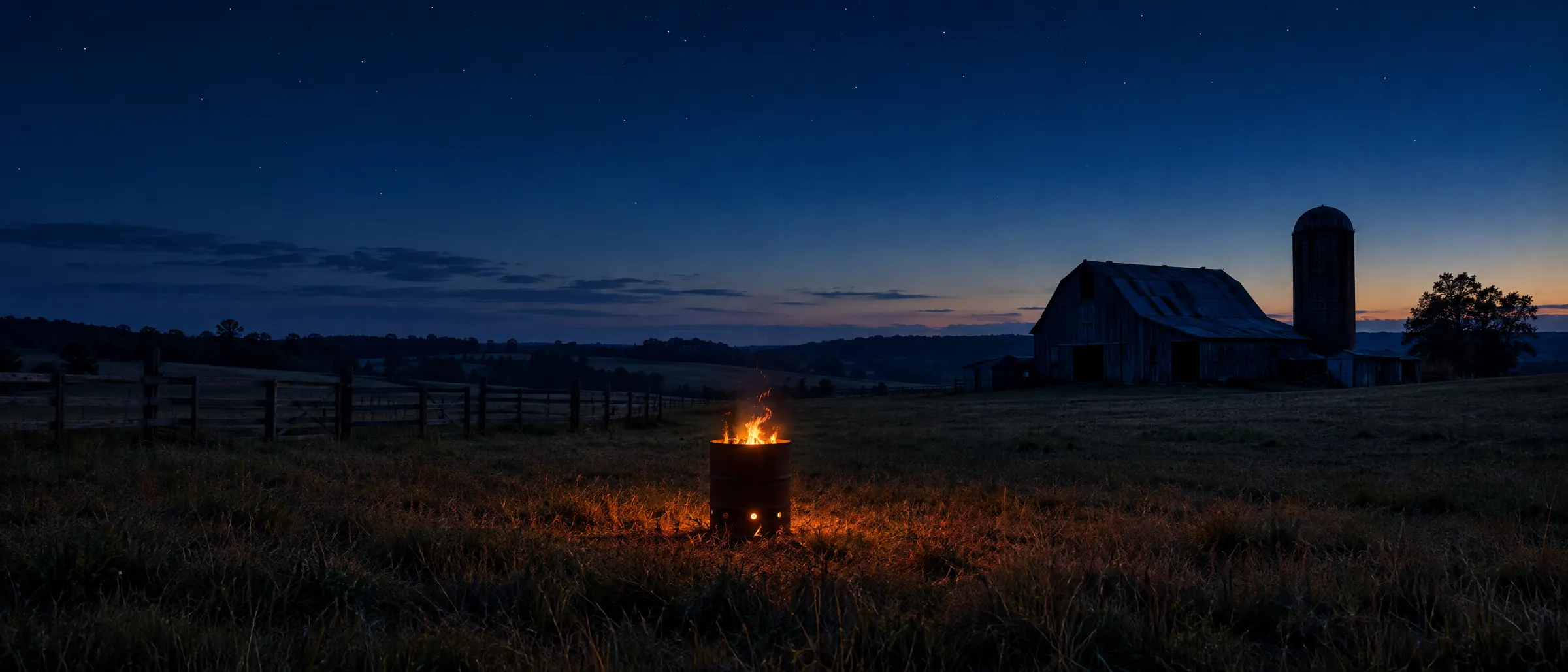 A farm at blue hour with a barn and silo in the distance and a small fire burning in a steel drum in the field.