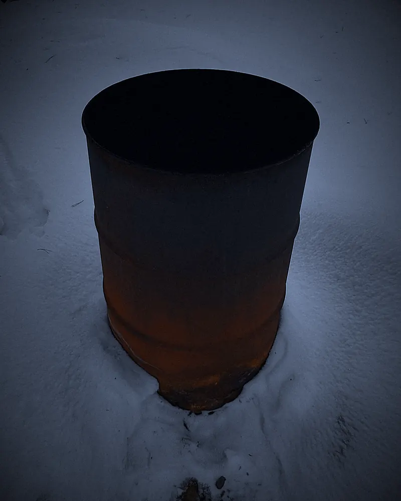 A rusted steel drum standing in snow at dusk.